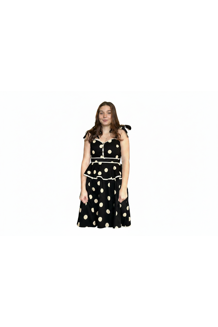 A woman modeling a black sleeveless dress with large beige polka dots, a sweetheart neckline, and tie details on the shoulders, set against a plain white background.