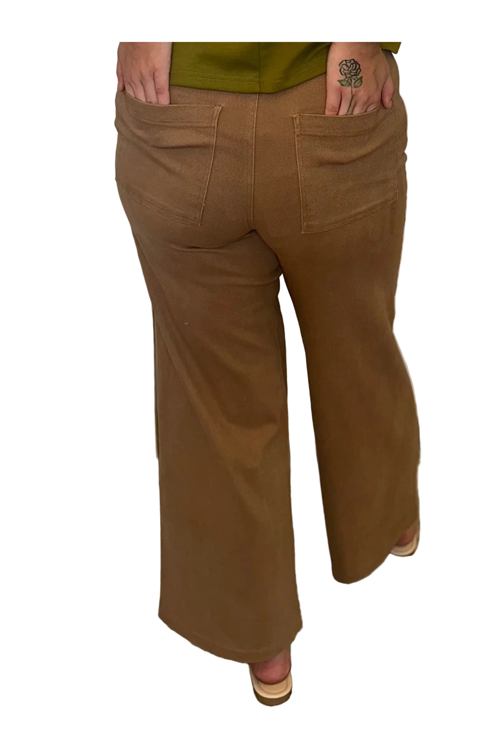 A woman stands with her back to the camera wearing an olive green short-sleeve top and brown wide-leg pants in front of white curtains.